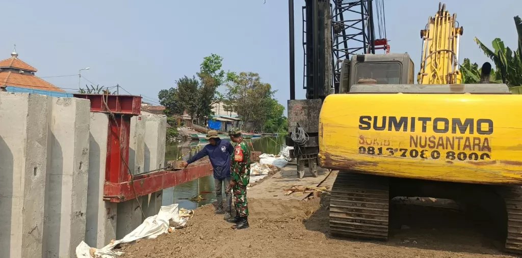 Babinsa Melaksanakan Pemantauan dan Monitoring Pengerjaan Pemancangan di Bantaran Sungai Menggunakan Alat Berat
